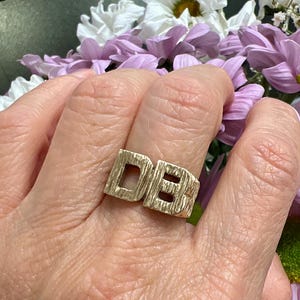 May include: A gold-coloured ring featuring the letters "DB" in a block letter design. The ring has a textured surface and is worn on a finger. The background includes purple and white flowers.