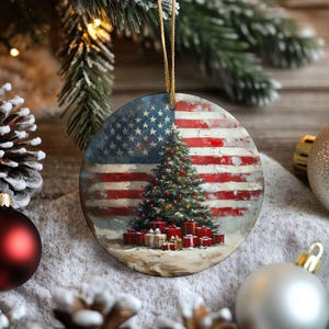 May include: Round Christmas ornament featuring the American flag design. The ornament has a painted image of a Christmas tree with red ornaments and wrapped gifts at the base. The ornament hangs from a gold string.