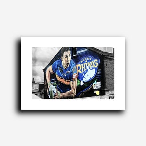 Puede incluir: Un mural colorido en un edificio de ladrillo representa a un jugador de rugby con una camiseta azul y amarilla sosteniendo una pelota. El mural incluye el texto "Leeds Rhinos" y un rinoceronte blanco.