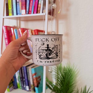 May include: White ceramic mug with black speckled design and the text "FUCK OFF I'm reading". The mug features a black illustration of a sword, dragon, and open book. The mug is held in front of a bookshelf.