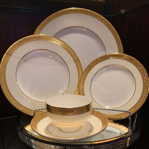 May include: A set of four white porcelain plates with gold trim and intricate designs. The plates are nested together, with the largest plate on the bottom and the smallest plate on top. A small white porcelain bowl with gold trim is also included.