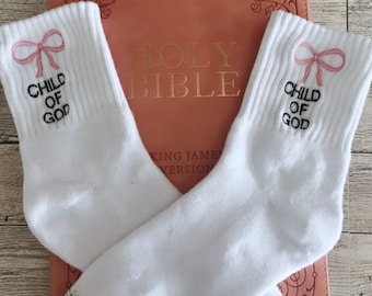 May include: A pair of white ankle socks with pink bows and the text "Child of God" embroidered on each sock. The socks are lying on top of a pink leather-bound Holy Bible.