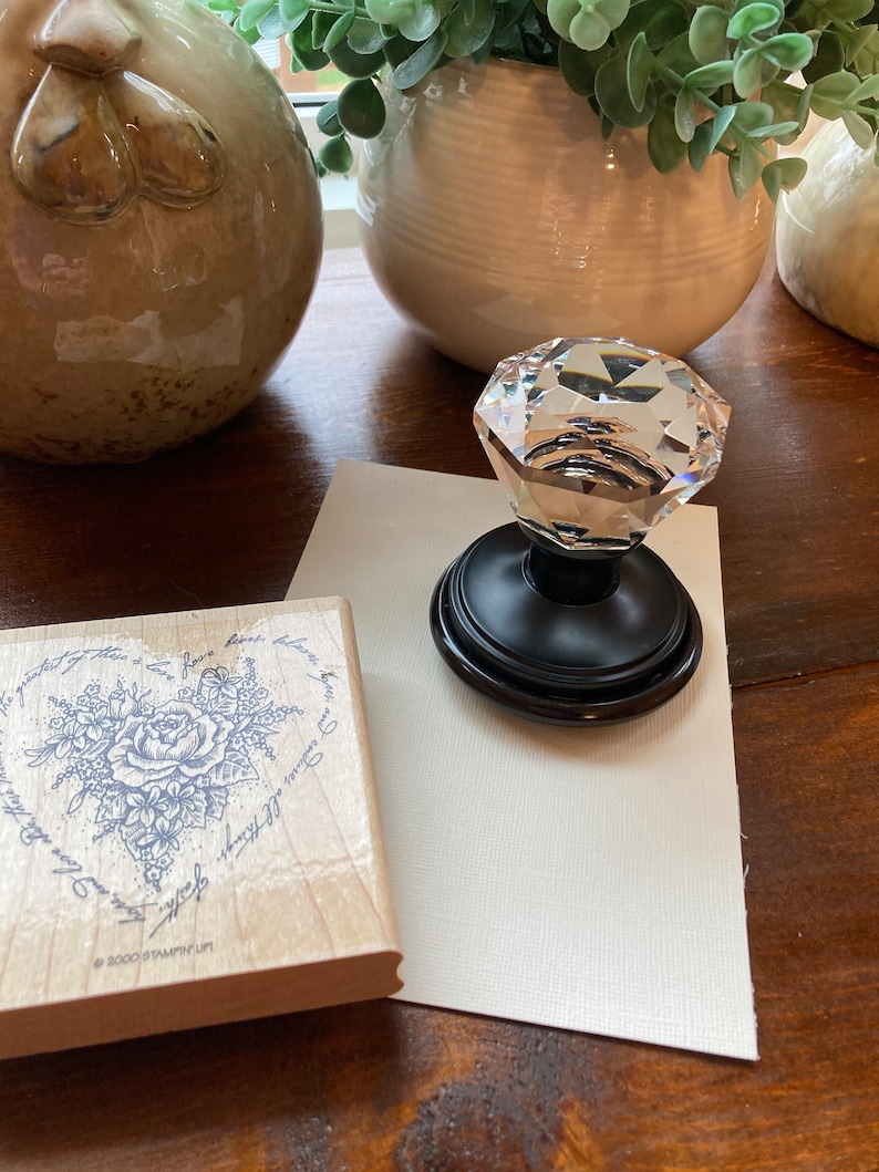 May include: A clear crystal knob with a black base sits on a white card next to a wooden stamp with a floral heart design. The stamp has text that reads "The greatest of these is love, love bears, and endures all things. Faith".