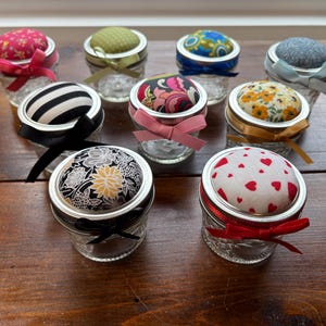 May include: A collection of eight small glass jars with metal lids topped with fabric pin cushions. Each lid features a unique fabric pattern, including floral, heart, and striped designs, and is accented with a coordinating satin ribbon bow.