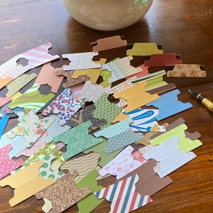 May include: A collection of colorful paper tabs with various patterns, including floral, striped, and geometric designs. The tabs are scattered on a wooden surface, with a decorative pen and a ceramic pot in the background. The tabs are ideal for organizing documents.