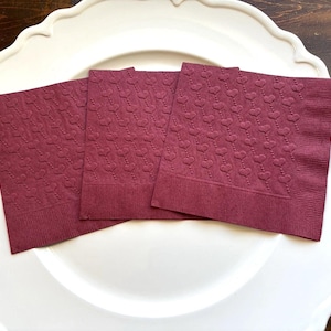 May include: Burgundy paper napkins with an embossed heart pattern, arranged on a white scalloped-edge plate. The napkins have a textured appearance and are folded neatly. The background is a dark wood surface.