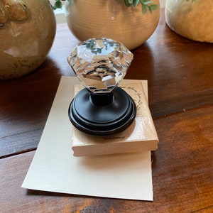 May include: A clear, faceted crystal knob with a black base sits atop a wooden stamp and a cream-colored card. The knob reflects light, and the stamp has text. Decorative items and greenery are in the background.