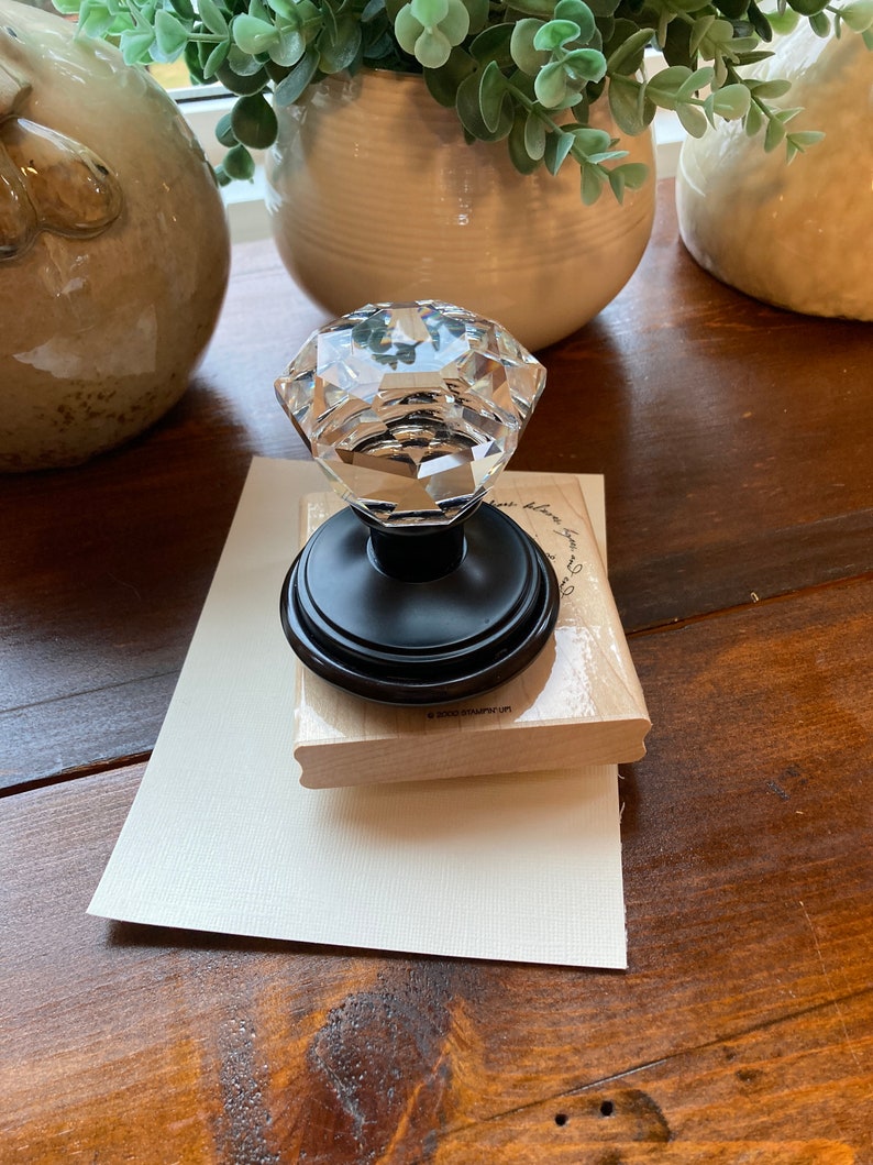 May include: A clear, faceted crystal knob with a black base sits atop a wooden stamp and a cream-colored card. The knob reflects light, and the stamp has text. Decorative items and greenery are in the background.