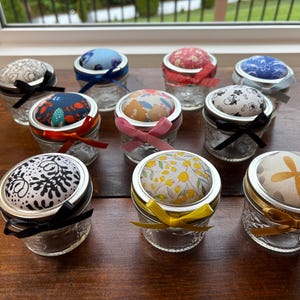 May include: A collection of nine small, clear glass jars with quilted patterns, each topped with a fabric-covered pin cushion lid. The lids feature various floral and abstract patterns in colors like blue, red, yellow, and black, accented with matching satin ribbons.