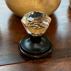 May include: A clear, faceted crystal knob with a black base. The knob is diamond-shaped and reflects light. The base is round and sits on a dark brown wooden surface. Decorative hardware.