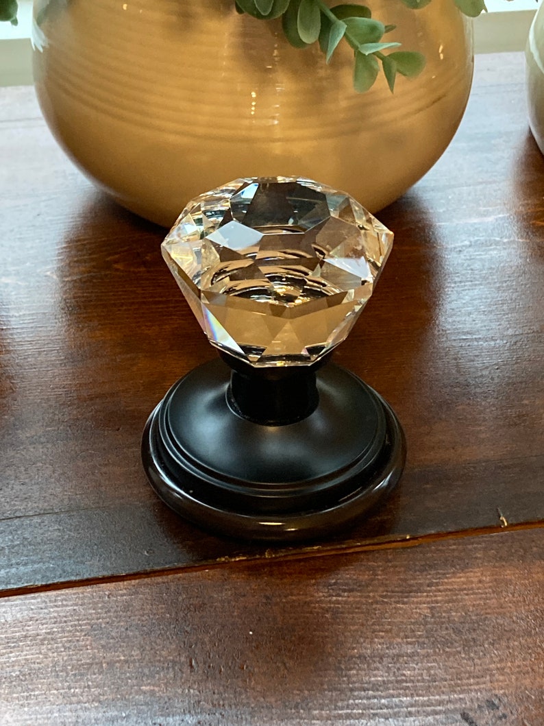 May include: A clear, faceted crystal knob with a black base. The knob is diamond-shaped and reflects light. The base is round and sits on a dark brown wooden surface. Decorative hardware.