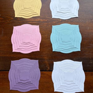 May include: Six layered paper cutouts in various pastel colors: yellow, white, pink, light blue, purple, and off-white. Each cutout has a unique wavy edge and a central square design, creating a 3D effect. The cutouts are arranged on a dark wood surface.