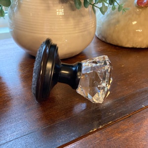 May include: A decorative cabinet knob with a faceted crystal handle and a dark brown base. The knob is set against a wooden surface, with a white ceramic pot and a decorative chicken in the background.