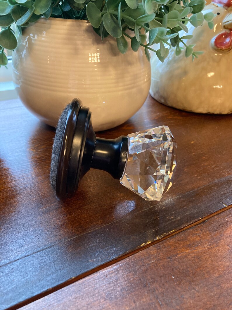 May include: A decorative cabinet knob with a faceted crystal handle and a dark brown base. The knob is set against a wooden surface, with a white ceramic pot and a decorative chicken in the background.