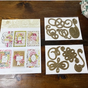 May include: A set of Anna Griffin metal cutting dies for paper crafting, including one instructional sheet displaying six finished card designs and two clear plastic sleeves containing various gold-toned, intricate scrollwork and flourish shapes.