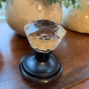 May include: A clear, faceted crystal door knob with a black base. The knob is diamond-shaped and reflects light. The base is round with a decorative edge. Home decor item.