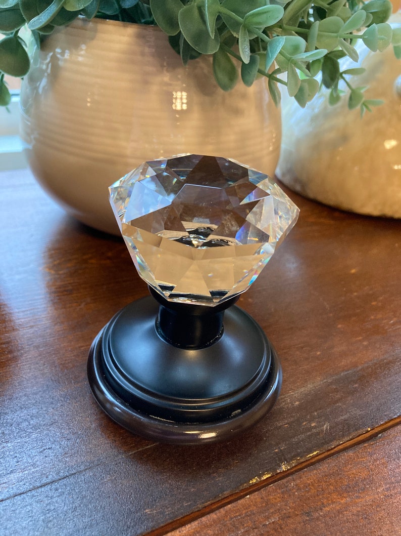 May include: A clear, faceted crystal door knob with a black base. The knob is diamond-shaped and reflects light. The base is round with a decorative edge. Home decor item.