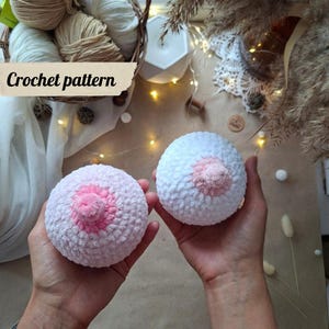 May include: Two round, plush, crocheted objects with a soft, fuzzy texture. Each piece is primarily white with a distinct, raised, light pink circular center. A beige label with the black text Crochet pattern is visible in the upper left corner.