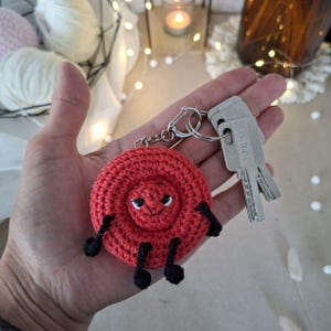 May include: A round, red crocheted keyring featuring a stitched face with black eyes and a small smile. The circular textile charm has four black yarn limbs attached to its sides and bottom, and it is connected to a silver metal keyring with two keys.