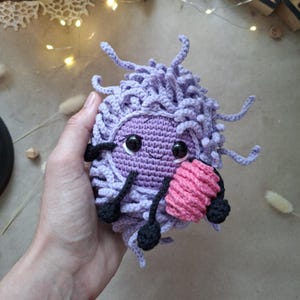 Peut inclure: Une peluche crochetée en forme de cellule avec un corps texturé violet clair, des yeux en plastique noir et une bouche souriante. Elle possède de fins bras et jambes noirs et tient un petit objet cylindrique rose dans l'une de ses mains noires.