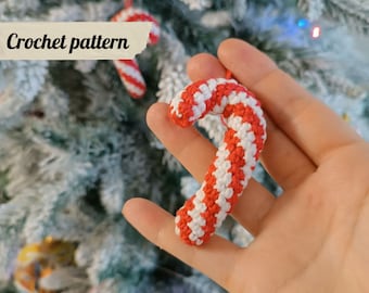 May include: A crocheted candy cane ornament in red and white, held in a hand against a background of a snowy Christmas tree.