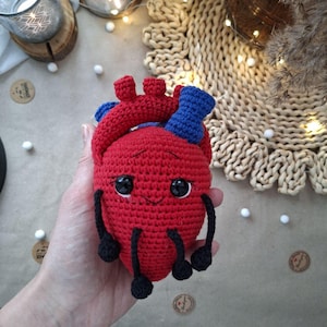 May include: A handmade, red crocheted heart-shaped plush toy with blue and black accents. The toy has large, black, googly eyes and a smiling mouth. The heart has small black legs and arms. The toy is held in a person's hand.