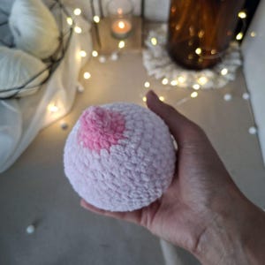 May include: A round, plush, crocheted object featuring a light pink, textured yarn body and a smaller, darker pink, raised, crocheted nipple-shaped center. The item is spherical in shape and crafted with a soft, fuzzy yarn material throughout.