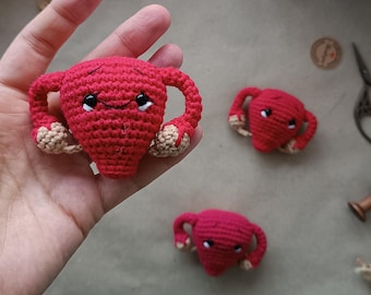 May include: Three crocheted uterus-shaped plushies in bright red yarn. Each piece features a smiling face with black and white embroidered eyes, a stitched mouth, and light tan yarn accents at the ends of the fallopian tubes. The texture is uniform.