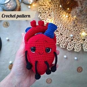 May include: A handmade red crochet heart with blue and black accents. The heart has black button eyes and a smiling face. The text "Crochet pattern" is in a banner above the heart.