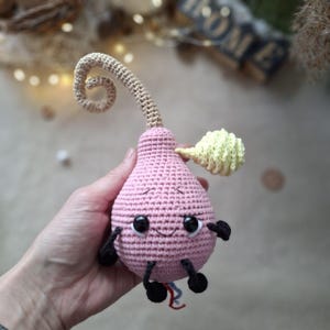 Könnte beinhalten: Ein rosa gehäkeltes Plüschtier in Tropfenform mit einem langen, lockigen beigen Stiel, einem kleinen gelben Blatt, schwarzen Kunststoffaugen, einem gestickten Lächeln und schwarzen Garnarmen und -beinen. Unten befindet sich ein kleiner rot-blauer Garnschwanz.