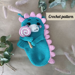 May include: Crochet pattern for a teal blue dinosaur holding a pink and white lollipop. The dinosaur has pink accents and a white face.
