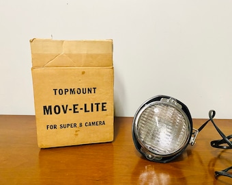 May include: A vintage Topmount Mov-E-Lite for Super 8 camera, featuring a round silver reflector light with a black housing and a black power cord, accompanied by its original tan cardboard box with black printed text on the front.