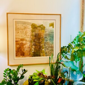 May include: A square framed print featuring a detailed architectural cityscape illustration. The artwork displays a dense urban scene with buildings and trees, transitioning from warm sepia tones on the left to cool blue-green hues on the right.