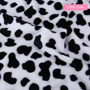May include: White fabric with a black cow print pattern. The fabric is soft and plush, with a fuzzy texture. The print is a classic black and white cow pattern, with large, irregular spots.