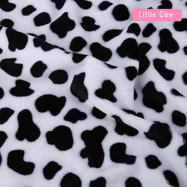 May include: White fabric with a black cow print pattern. The fabric is soft and plush, with a fuzzy texture. The print is a classic black and white cow pattern, with large, irregular spots.