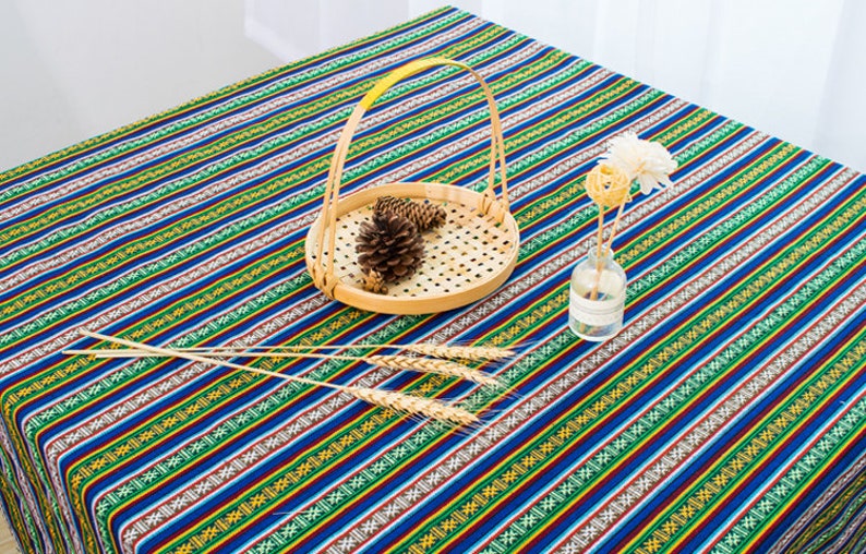 May include: A blue and green striped tablecloth with a woven basket and dried flowers in a glass vase. The tablecloth has a repeating pattern of small geometric shapes.