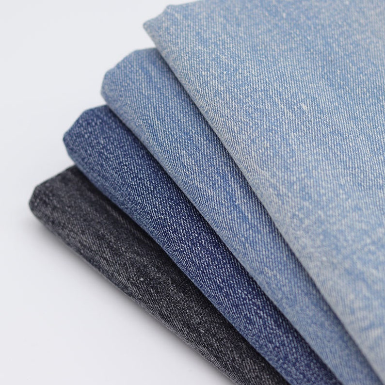 Puede incluir: Una pila de cinco muestras de tela vaquera en varios tonos de azul y negro. La tela tiene una textura sutil y una apariencia ligeramente descolorida.