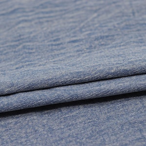Puede incluir: Primer plano de una tela vaquera azul claro con un sutil tejido blanco. La tela est&aacute; doblada por la mitad, mostrando la textura y el tejido del material.