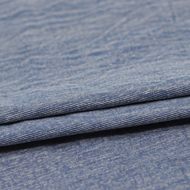 Puede incluir: Primer plano de una tela vaquera azul claro con un sutil tejido blanco. La tela est&aacute; doblada por la mitad, mostrando la textura y el tejido del material.