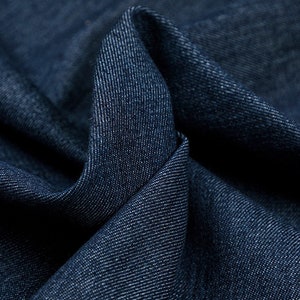 May include: Close-up of a dark blue denim fabric with a textured weave. The fabric is folded and draped, showing the soft texture and deep color.