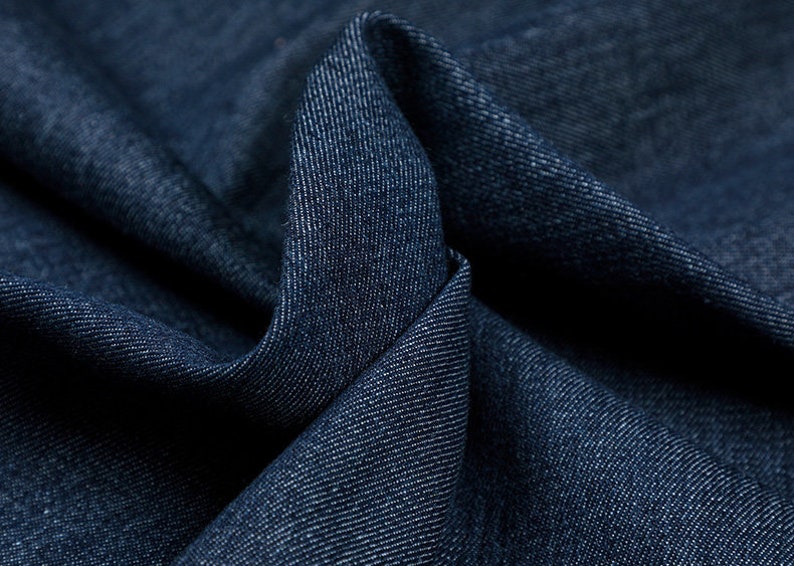 May include: Close-up of a dark blue denim fabric with a textured weave. The fabric is folded and draped, showing the soft texture and deep color.
