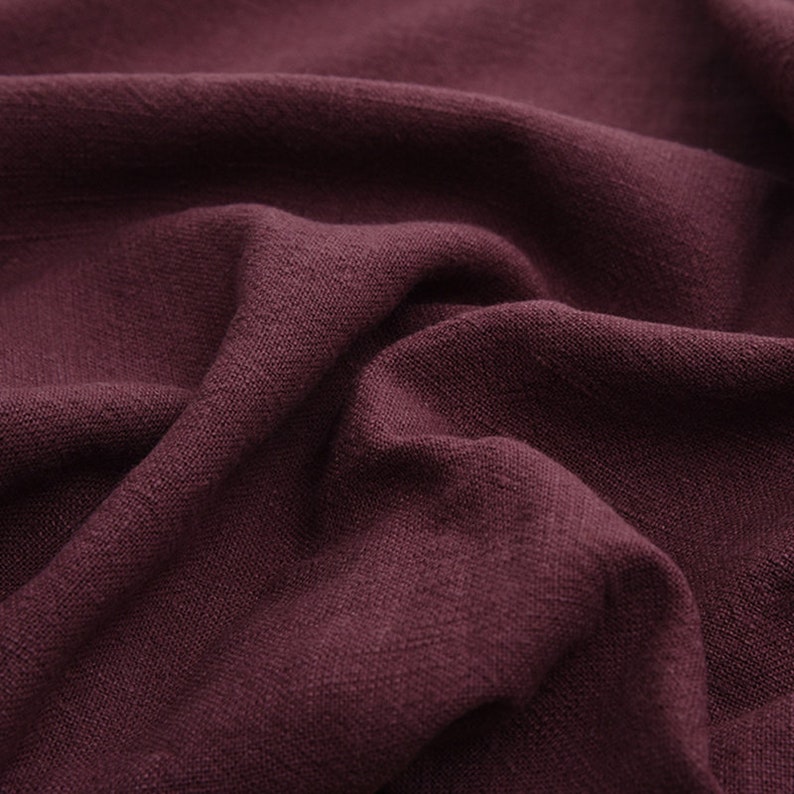 May include: Close-up of a deep burgundy fabric with a textured, linen-like weave. The fabric is folded and draped, showcasing its soft, drapey texture.