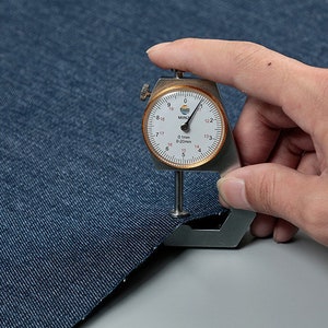 May include: A close-up of a blue denim fabric being measured with a fabric thickness gauge. The gauge reads 0.1mm or 0.020mm.