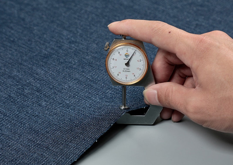 May include: A close-up of a blue denim fabric being measured with a fabric thickness gauge. The gauge reads 0.1mm or 0.020mm.