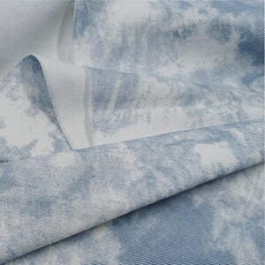 Pu&ograve; includere: Primo piano di un tessuto tie-dye blu chiaro e bianco con una texture sottile. Il tessuto &egrave; piegato a met&agrave;, mostrando il motivo su entrambi i lati.