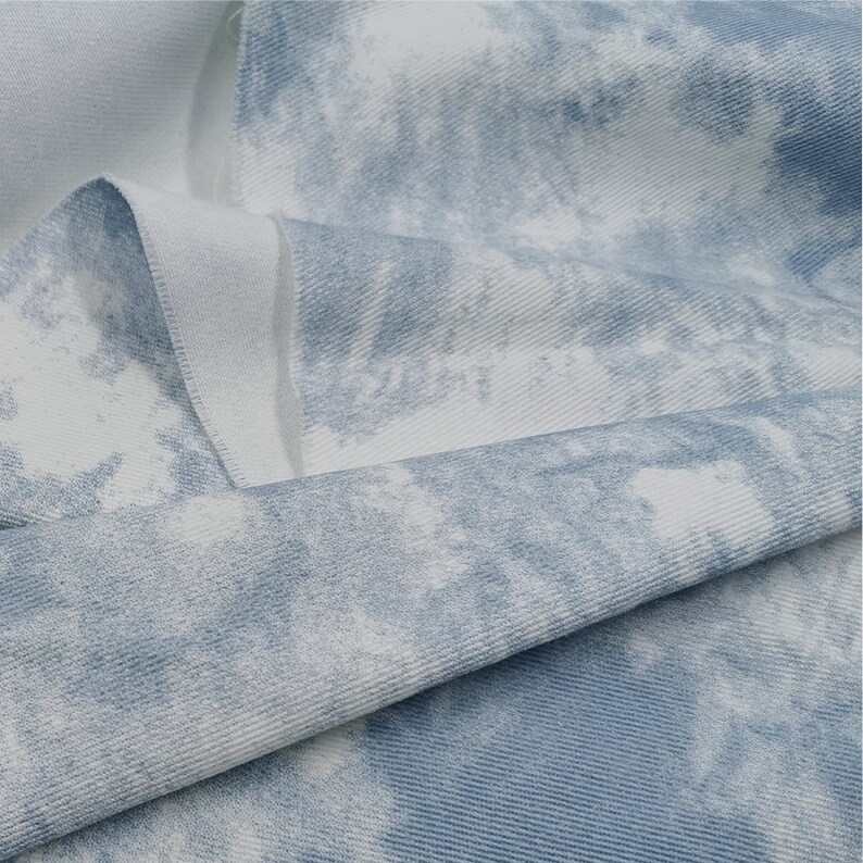 Pu&ograve; includere: Primo piano di un tessuto tie-dye blu chiaro e bianco con una texture sottile. Il tessuto &egrave; piegato a met&agrave;, mostrando il motivo su entrambi i lati.