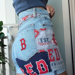 Könnte beinhalten: Hellblaue Jeansshorts mit Aufnähern des Boston Red Sox in Rot und Weiß. Die Shorts haben einen Used-Look, Sternverzierungen und einen marineblauen Aufnäher. Die Aufnäher zeigen das Logo und die Schrift der Mannschaft.
