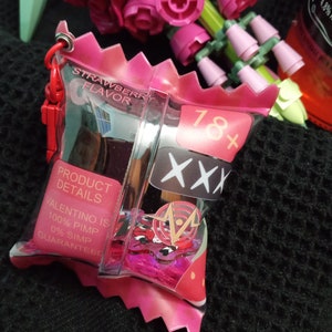 Puede incluir: Una bolsa de pl&aacute;stico rosa con cierre de cremallera y una etiqueta de sabor a fresa. La bolsa contiene un peque&ntilde;o recipiente de pl&aacute;stico transparente con un dise&ntilde;o rosa y rojo. El recipiente tiene el texto "18+ XXX" y "VALENTINO IS 100% PIMP 0% SIMP GUARANTEED" impreso en &eacute;l.