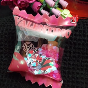 Puede incluir: Una bolsa de pl&aacute;stico rosa con la palabra "VALENTINO" impresa en ella. La bolsa contiene una peque&ntilde;a botella de l&iacute;quido y corazones de purpurina rosa.