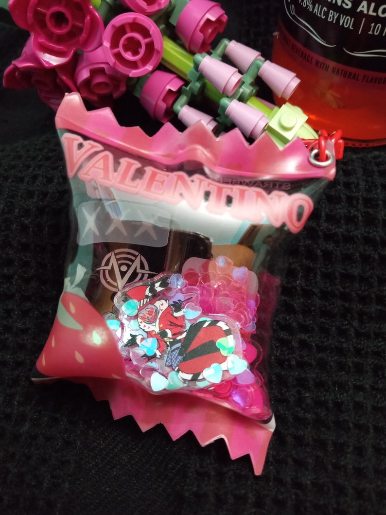 Puede incluir: Una bolsa de pl&aacute;stico rosa con la palabra "VALENTINO" impresa en ella. La bolsa contiene una peque&ntilde;a botella de l&iacute;quido y corazones de purpurina rosa.